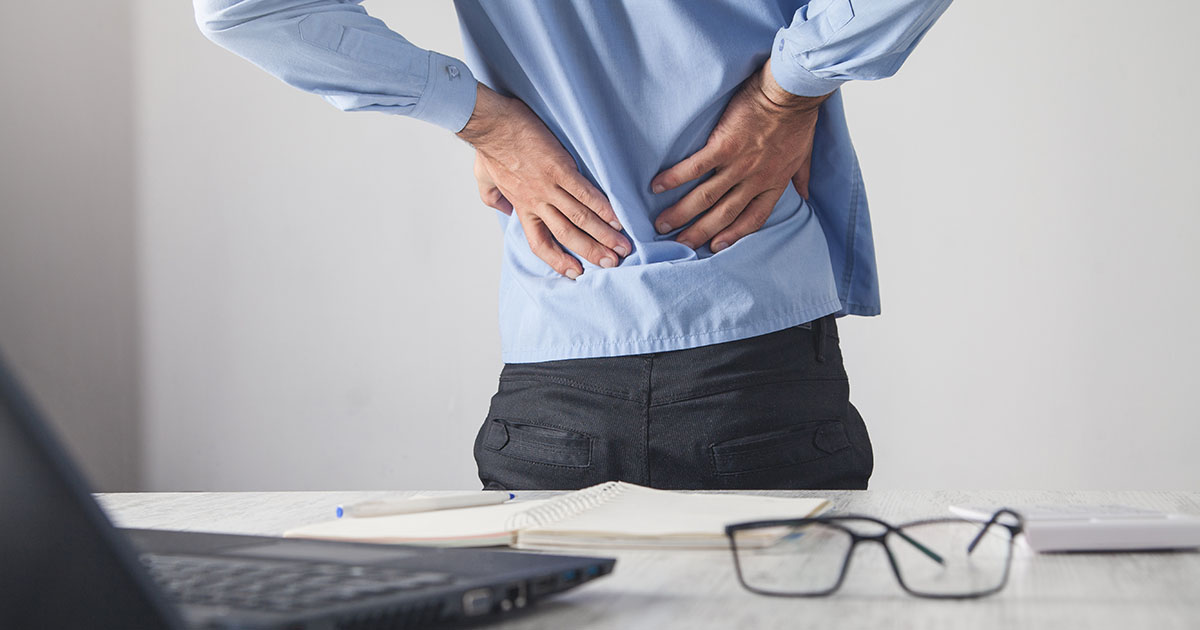 Man in a blue shirt standing at a desk, both hands pressed into his lower back in visible pain, laptop and notepad on the desk in front of him