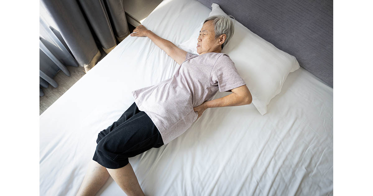 Older woman lying on her side in bed, pressing her hand into her lower back in pain, illustrating back pain that worsens after rest