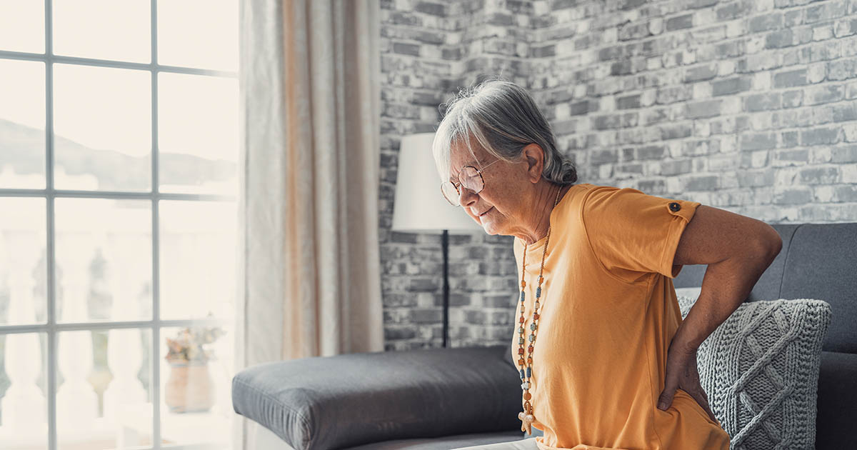 Older woman sitting on the edge of a couch and holding her lower back in pain, illustrating recurring back pain that returns after temporary treatment