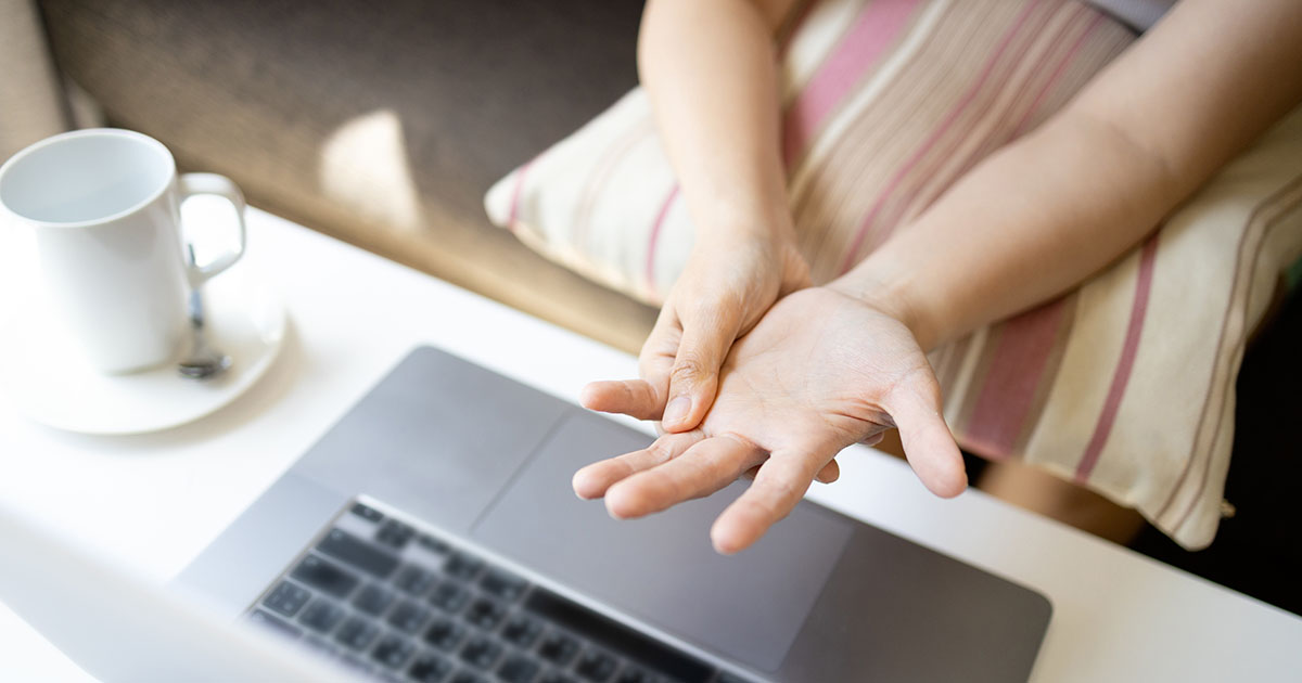 Person sitting at a laptop massaging one palm and wrist with the other hand, a common sign of wrist pain from prolonged typing